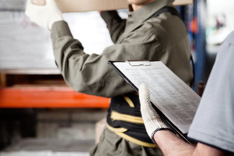 Midsection of foreman loading cardboard box with supervisor holding clipboard at warehouse. Midsection of foreman loading cardboard box with supervisor holding clipboard at warehouse