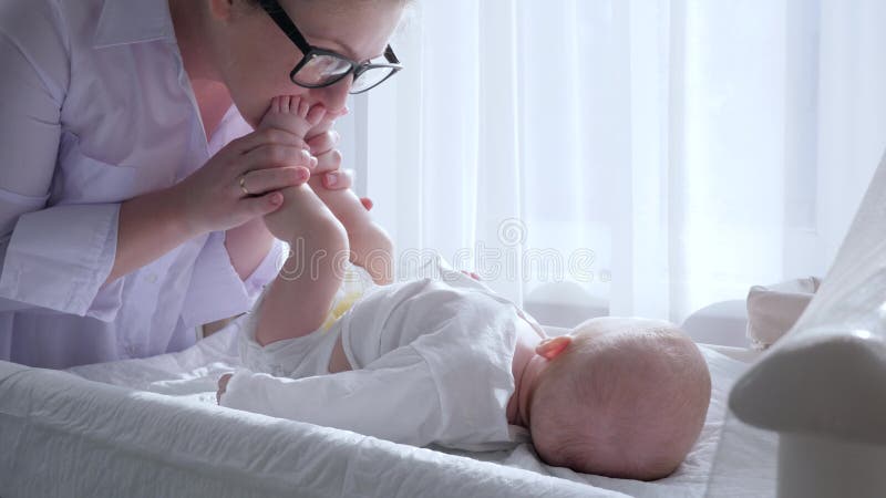 Материнская нежность, мама одевает ребенка и целует ногу в светлой комнате против занавесов