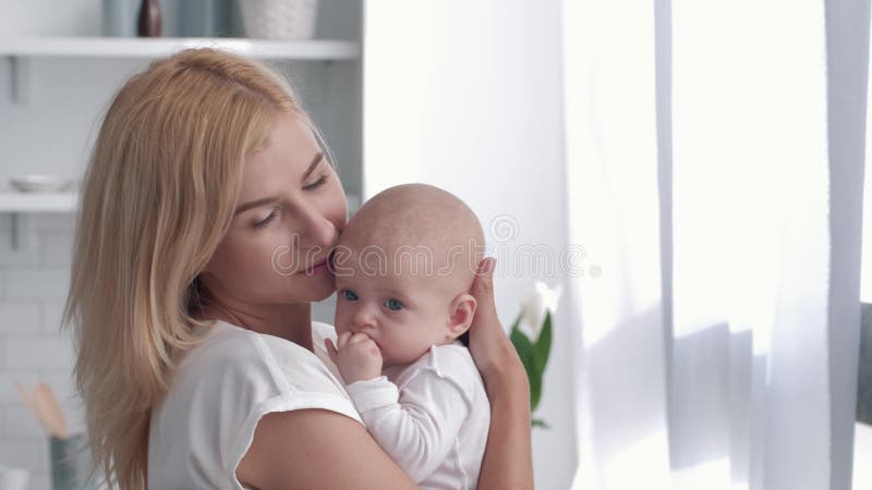 Материнская нежность, молодой родитель обнимает и целует newborn ребенка на кухне дома