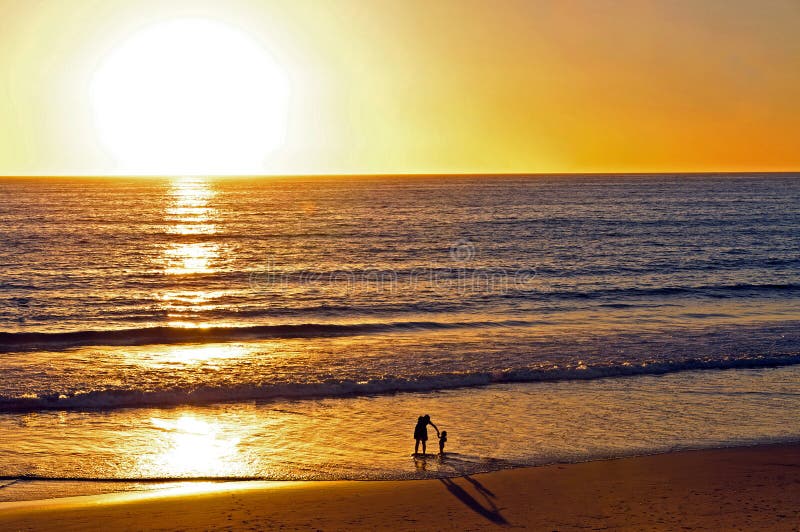 Мать и ребенок играя на пляже на заходе солнца