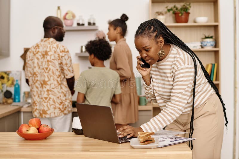 Black woman eating breakfast sandwich, talking on phone and answering e-mails when her family cooking in background. Black woman eating breakfast sandwich, talking on phone and answering e-mails when her family cooking in background