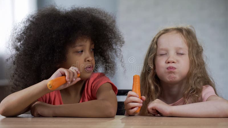 Меньшие довольно multiracial девушки сидя на таблице и есть морковь с аппетитом стоковая фотография rf