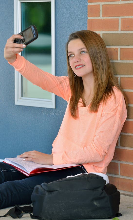 Adorable young girl, with braces, sitting in front of school building with book open on lap but taking photo of self with cell phone camera. Adorable young girl, with braces, sitting in front of school building with book open on lap but taking photo of self with cell phone camera