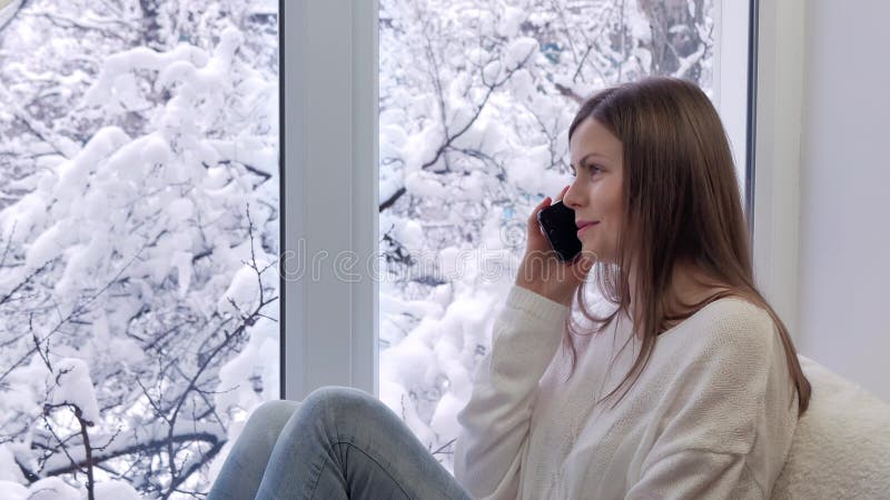 Милая девушка сидя на windowsill говоря на smartphone внешняя зима