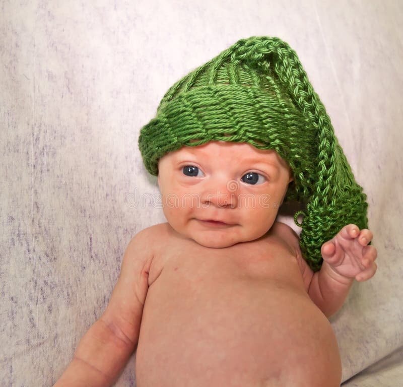 Sweet newborn baby girl wearing a knit stocking hat that makes her look like a cute little elf. Infant is approximately two months old. Blue eyes. White and purple fabric background. Sweet newborn baby girl wearing a knit stocking hat that makes her look like a cute little elf. Infant is approximately two months old. Blue eyes. White and purple fabric background.