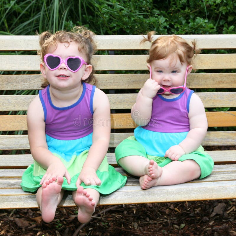 Adorable image of 2 little girls sitting on a park bench in sun dresses and sunglasses. Adorable image of 2 little girls sitting on a park bench in sun dresses and sunglasses