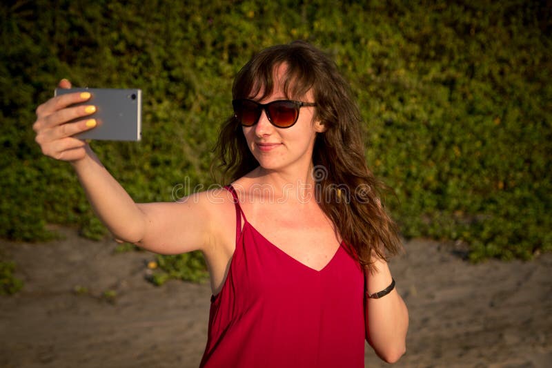 Молодая женщина в красном платье делая фото selfie на пляже Северный g стоковое изображение rf Молодая женщина в красном платье делая фото selfie на пляже Северный g стоковое изображение rf