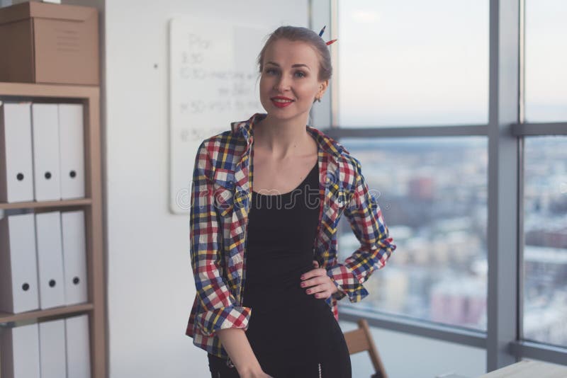 Молодая женщина нося вскользь яркую рубашку стоя в светлом просторном офисе, ее рабочем месте, подготавливает на день работы, усм стоковые изображения rf Молодая женщина нося вскользь яркую рубашку стоя в светлом просторном офисе, ее рабочем месте, подготавливает на день работы, усм стоковые изображения rf