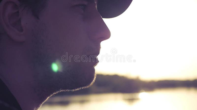Молодой красивый человек сидит в профиле на береге озера на предпосылке захода солнца Конец-вверх Hd 1080p замедленного движения
