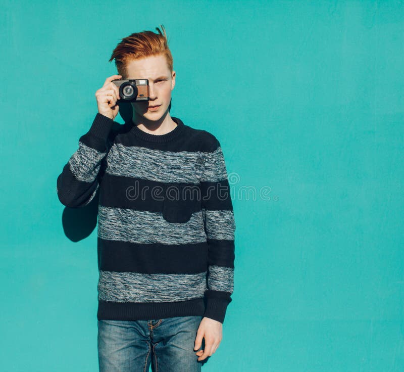 Молодой человек redhead в свитере и джинсах стоя рядом с стеной бирюзы и принимая фото винтажную камеру теплое лето солнечный ден стоковые изображения rf Молодой человек redhead в свитере и джинсах стоя рядом с стеной бирюзы и принимая фото винтажную камеру теплое лето солнечный ден стоковые изображения rf