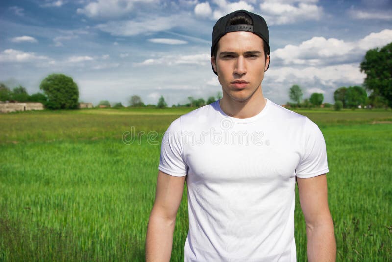 Handsome fit young man at countryside, in front of field or grassland, wearing white t-shirt and cap, looking at camera. Handsome fit young man at countryside, in front of field or grassland, wearing white t-shirt and cap, looking at camera