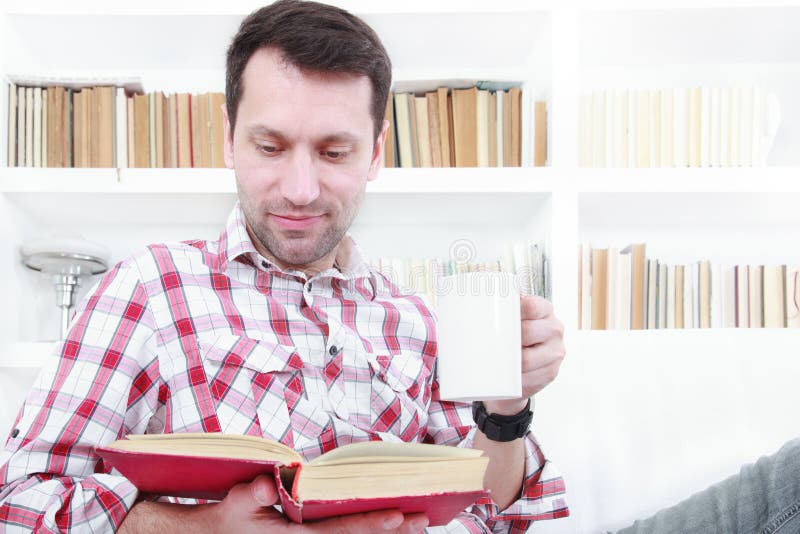 Молодой человек ослабляя на книге чтения кресла и наслаждаясь кофе стоковая фотография rf Молодой человек ослабляя на книге чтения кресла и наслаждаясь кофе стоковая фотография rf