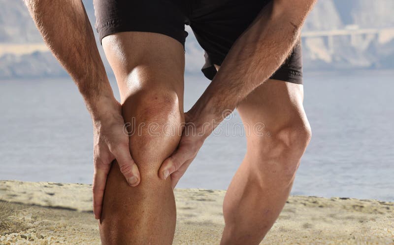 Young sport man with strong athletic legs holding knee with his hands in pain after suffering muscle injury during a running workout beach training in muscular or ligament wound. Young sport man with strong athletic legs holding knee with his hands in pain after suffering muscle injury during a running workout beach training in muscular or ligament wound