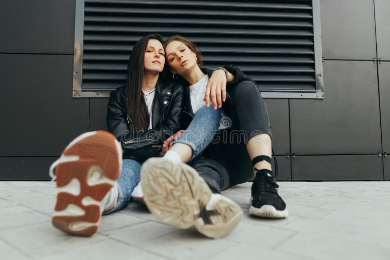 Fashionable photo of two stylish girls in casual clothes wearing leather jackets and sneakers,sitting on the ground against a dark wall and posing in camera.Fashion portrait 2 girlfriends on street. Fashionable photo of two stylish girls in casual clothes wearing leather jackets and sneakers,sitting on the ground against a dark wall and posing in camera.Fashion portrait 2 girlfriends on street