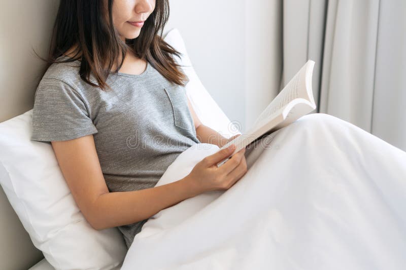 молодая азиатка в пижаме читает книгу, сидя на кровати утром дома. отдых и отдых стоковые изображения rf