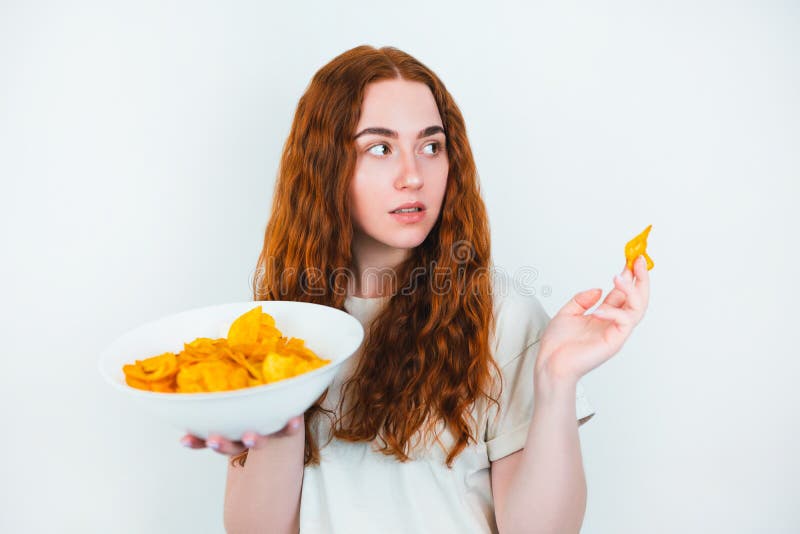 Молодая женщина, есть redheaded хрустящие картофельные чипсы выглядит счастливой на изолированной белой концепции высококалорийной стоковое фото