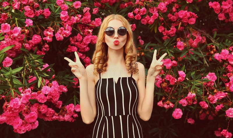 Fashion portrait beautiful young woman blowing red lips sending sweet air kiss over pink flowers background. Fashion portrait beautiful young woman blowing red lips sending sweet air kiss over pink flowers background