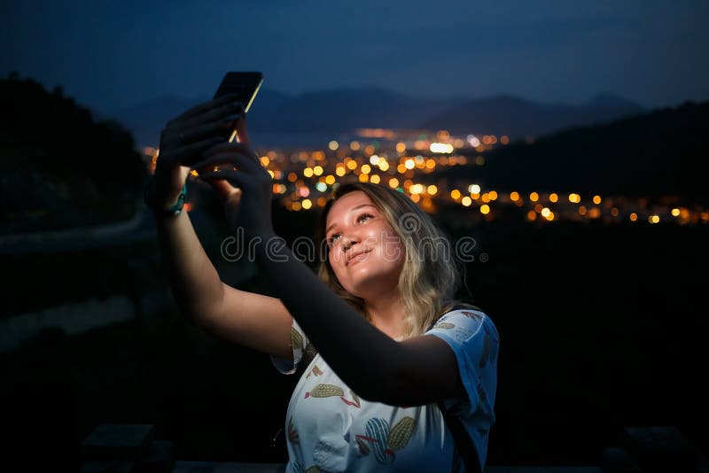 Молодая женщина принимая selfie поверх холма наблюдающ видом на город ночи Хороший свет от экрана стоковое изображение rf Молодая женщина принимая selfie поверх холма наблюдающ видом на город ночи Хороший свет от экрана стоковое изображение rf