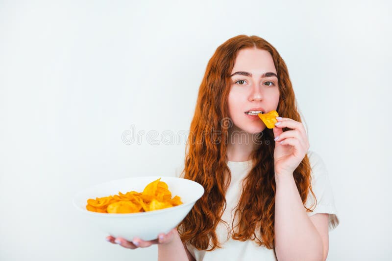 Молодая женщина с укусами redheaded хрустящие картофельные чипсы выглядит счастливой на изолированной белой концепции высококалори стоковая фотография rf
