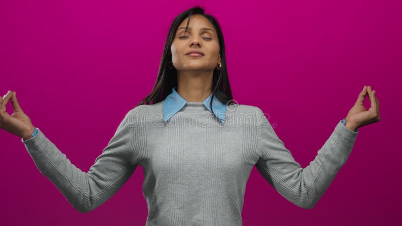 Молодая испаноязычная женщина медитирует с закрытыми глазами на ярком розовом фоне, воплощая спокойствие и безмятежность в совреме сток-видео