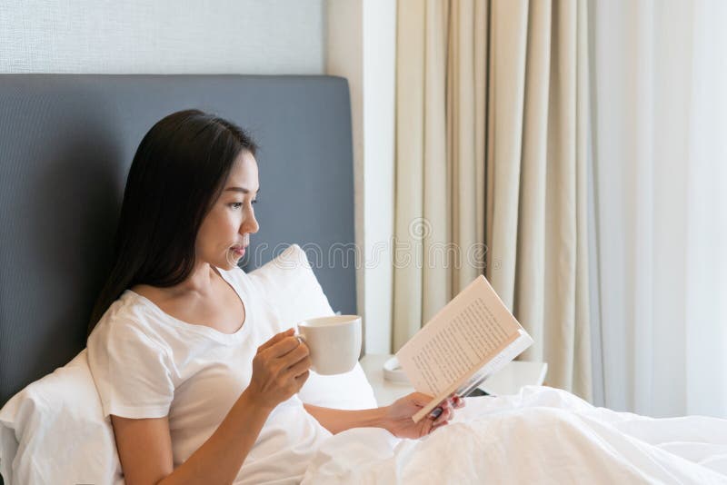 молодая красивая азиатка в белой футболке пьёт кофе, читая книгу, сидящую на кровати утром. копировать стоковые фотографии rf