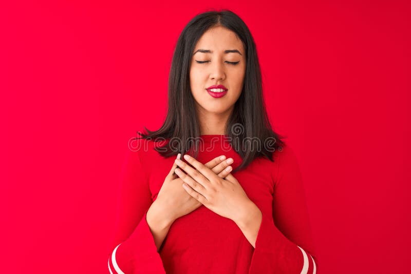 Young beautiful chinese woman wearing casual dress standing over isolated red background smiling with hands on chest with closed eyes and grateful gesture on face. Health concept. Young beautiful chinese woman wearing casual dress standing over isolated red background smiling with hands on chest with closed eyes and grateful gesture on face. Health concept