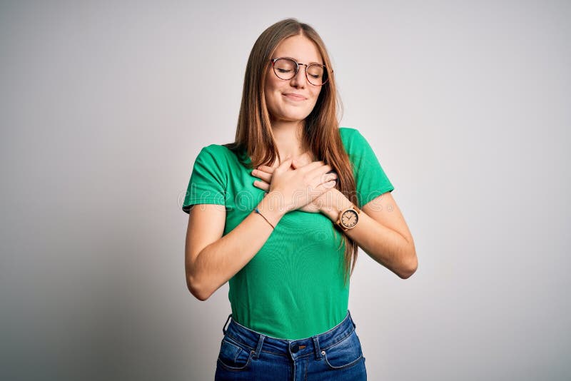 Young beautiful redhead woman wearing casual green t-shirt and glasses over white background smiling with hands on chest with closed eyes and grateful gesture on face. Health concept. Young beautiful redhead woman wearing casual green t-shirt and glasses over white background smiling with hands on chest with closed eyes and grateful gesture on face. Health concept