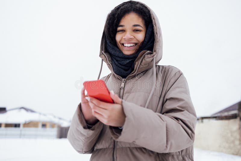 Young enchanting african woman in warm casual clothes holds phone. Joyful mixed race girl with bright spring flowers sending message with mobile. Winter snow background. Young enchanting african woman in warm casual clothes holds phone. Joyful mixed race girl with bright spring flowers sending message with mobile. Winter snow background