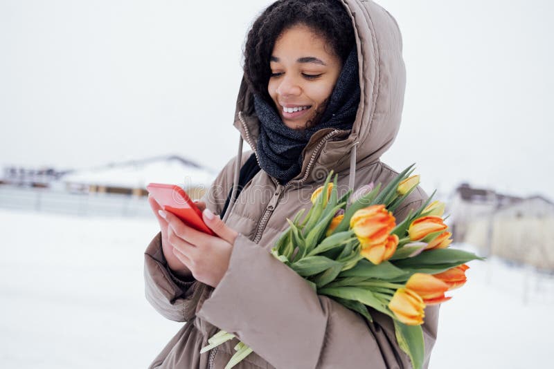 Young enchanting african woman in warm casual clothes holds phone and bouquet of orange tulips. Joyful mixed race girl with bright spring flowers sending message with mobile. Winter snow background. Young enchanting african woman in warm casual clothes holds phone and bouquet of orange tulips. Joyful mixed race girl with bright spring flowers sending message with mobile. Winter snow background