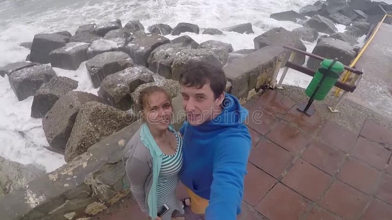 Молодая пара, человек и женщина стоят на пристани и делают selfie на пляже, волны ломают против больших камней медленно