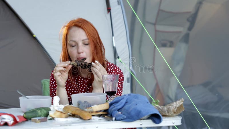 молодая рыжая женщина ест мясной стейк в кемпинге у палатки в сосновом лесу. видеоматериал