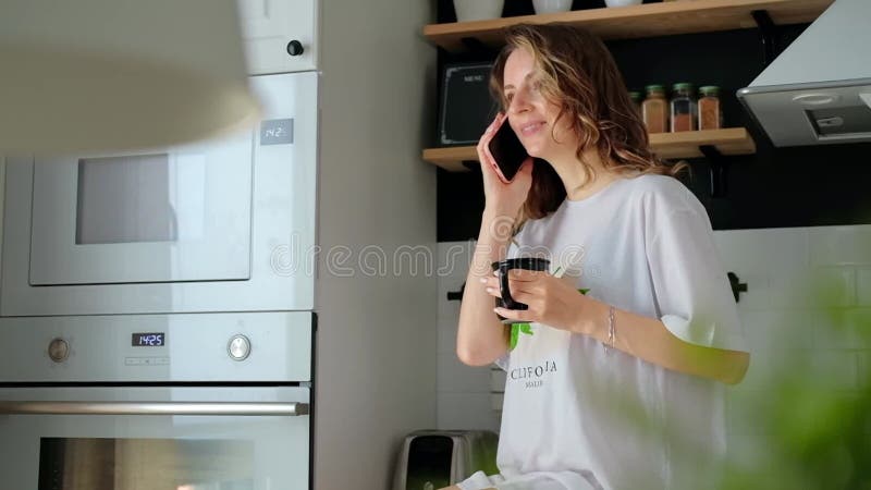 молодая симпатичная женщина пьет кофе сидя у себя дома на кухне видеоматериал