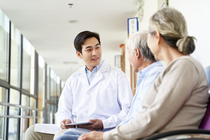 Молодой азиатский врач разговаривает со пожилой парой пациентов в больничном коридоре стоковые изображения