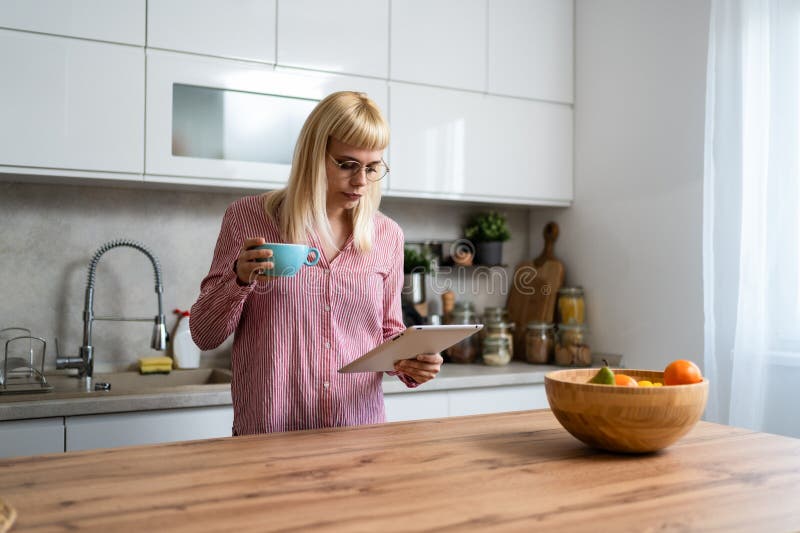 Young business woman, small company owner, startup businessperson expert marketing and product strategy developer, drinking first coffee in the kitchen using digital tablet to see statistics and mails. Young business woman, small company owner, startup businessperson expert marketing and product strategy developer, drinking first coffee in the kitchen using digital tablet to see statistics and mails