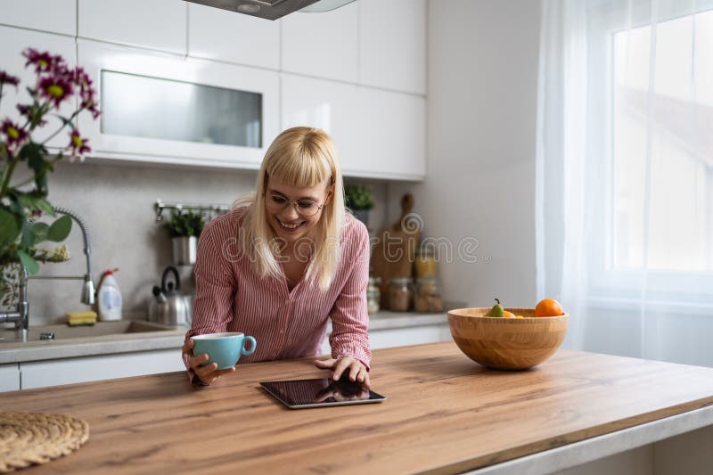 Young business woman, small company owner, startup businessperson expert marketing and product strategy developer, drinking first coffee in the kitchen using digital tablet to see statistics and mails. Young business woman, small company owner, startup businessperson expert marketing and product strategy developer, drinking first coffee in the kitchen using digital tablet to see statistics and mails