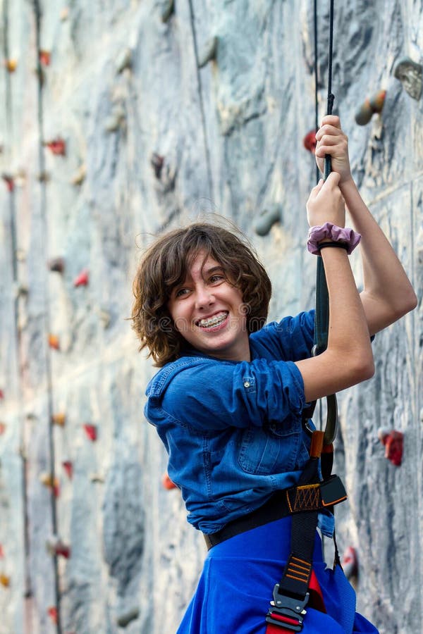 A young girl turns to smile at friends as she repels down a play rock climbing wall. She has braces and a snaggle tooth. There is a hair tie on her wrist. A young girl turns to smile at friends as she repels down a play rock climbing wall. She has braces and a snaggle tooth. There is a hair tie on her wrist.