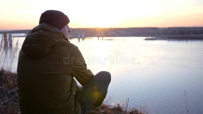 Молодой человек в куртке и шляпе сидит на речном береге около моста и восхищает красивый заход солнца движение медленное