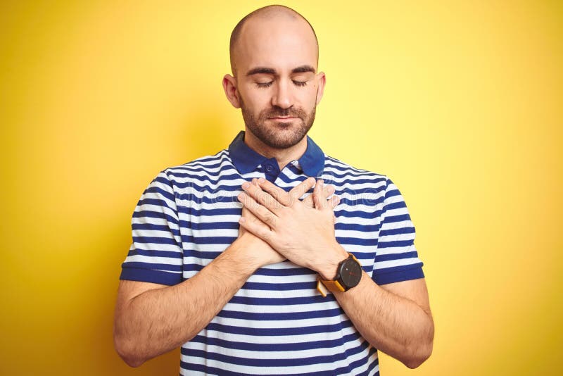 Young bald man with beard wearing casual striped blue t-shirt over yellow isolated background smiling with hands on chest with closed eyes and grateful gesture on face. Health concept. Young bald man with beard wearing casual striped blue t-shirt over yellow isolated background smiling with hands on chest with closed eyes and grateful gesture on face. Health concept