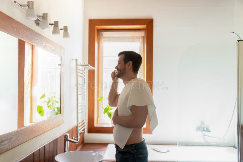 Millennial Caucasian man look in mirror do daily facial morning routine after shower in modern bright bathroom, young male clean brush teeth with toothbrush in bath, oral hygiene concept. Millennial Caucasian man look in mirror do daily facial morning routine after shower in modern bright bathroom, young male clean brush teeth with toothbrush in bath, oral hygiene concept