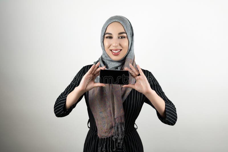 Молодые красивые усмехаясь показ головного платка hijab тюрбана мусульманской женщины нося и смартфон удержания в ее руках изолир стоковые изображения rf Молодые красивые усмехаясь показ головного платка hijab тюрбана мусульманской женщины нося и смартфон удержания в ее руках изолир стоковые изображения rf
