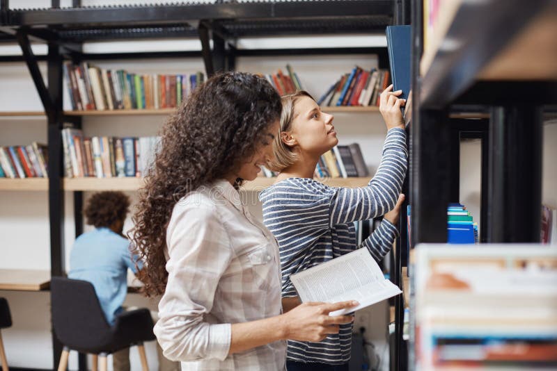 2 молодых жизнерадостных студентки в вскользь одеждах стоя близко книжные полки в университетской библиотеке смотря до конца стоковые фотографии rf 2 молодых жизнерадостных студентки в вскользь одеждах стоя близко книжные полки в университетской библиотеке смотря до конца стоковые фотографии rf