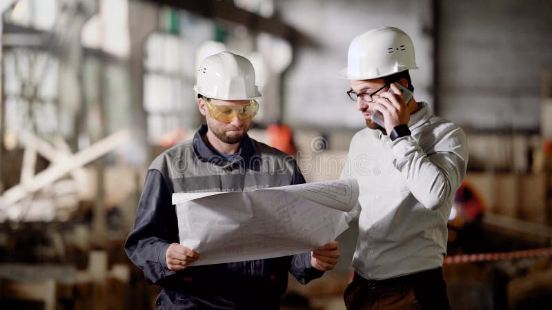 2 мужских инженера стоя в строительной площадке и держа план строительства Дизайнер говоря на телефоне