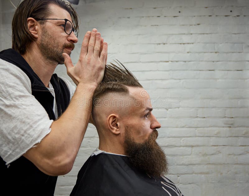 Мужской парикмахер делает стиль причесок mohawk используя клипер взрослого человека с бородой стоковые изображения rf Мужской парикмахер делает стиль причесок mohawk используя клипер взрослого человека с бородой стоковые изображения rf