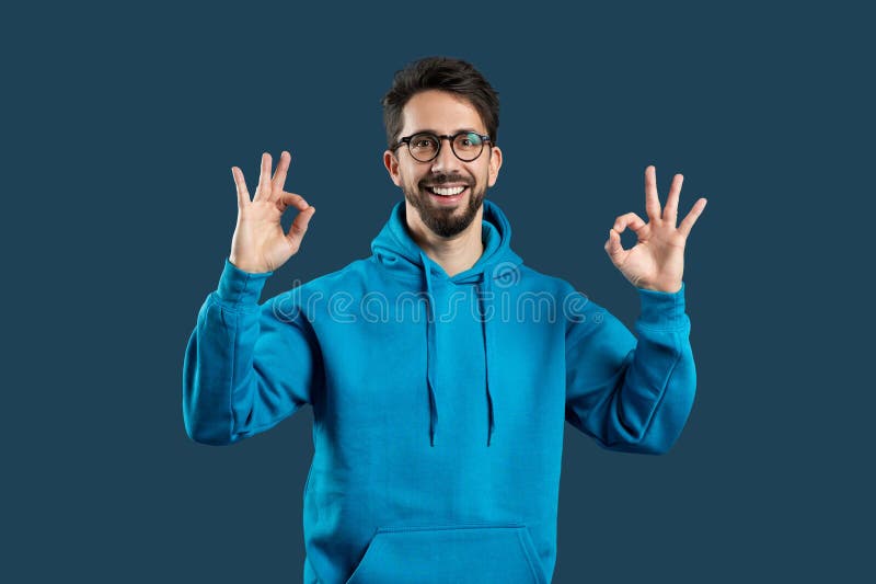 A cheerful man with a beard and glasses is wearing a bright blue hoodie and making the OK sign with both hands, suggesting positivity and approval. He is standing against a monochrome blue background. A cheerful man with a beard and glasses is wearing a bright blue hoodie and making the OK sign with both hands, suggesting positivity and approval. He is standing against a monochrome blue background