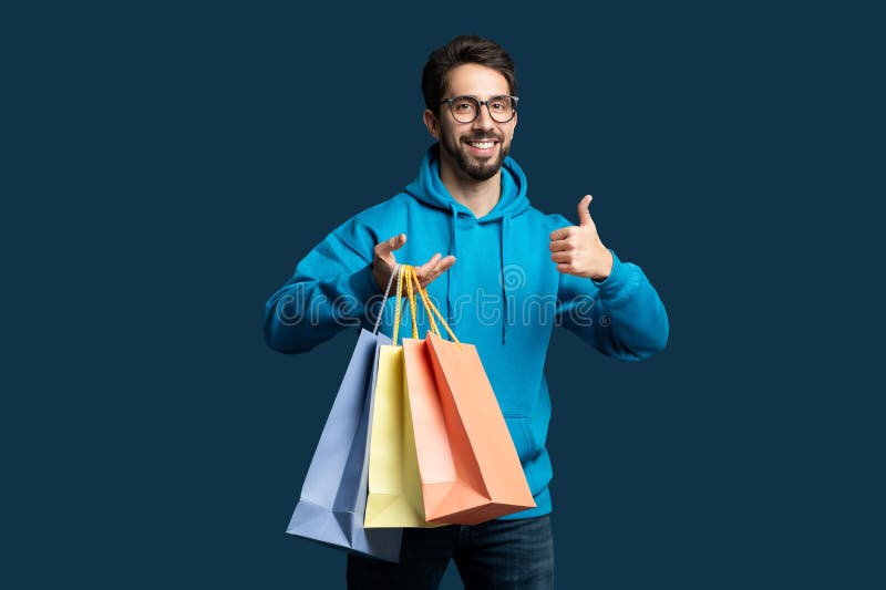 A man wearing a blue hoodie is standing while holding shopping bags in his hands. He is smiling and giving a thumbs up gesture, expressing satisfaction with his purchases. A man wearing a blue hoodie is standing while holding shopping bags in his hands. He is smiling and giving a thumbs up gesture, expressing satisfaction with his purchases.