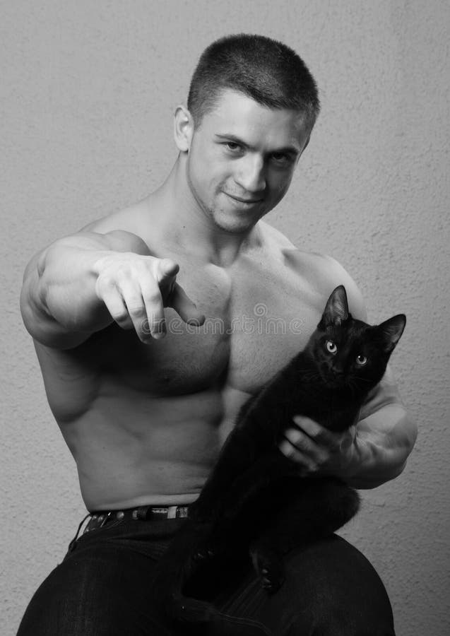 Black and white studio portrait of a muscular young bodybuilder pointing toward the viewer, holding a black cat in his other hand. Black and white studio portrait of a muscular young bodybuilder pointing toward the viewer, holding a black cat in his other hand.