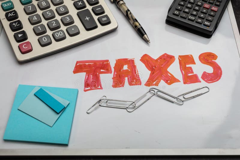The word TAXES in red on a dry erase board office supplies calculators posted notes and pen accounting finance industry. The word TAXES in red on a dry erase board office supplies calculators posted notes and pen accounting finance industry