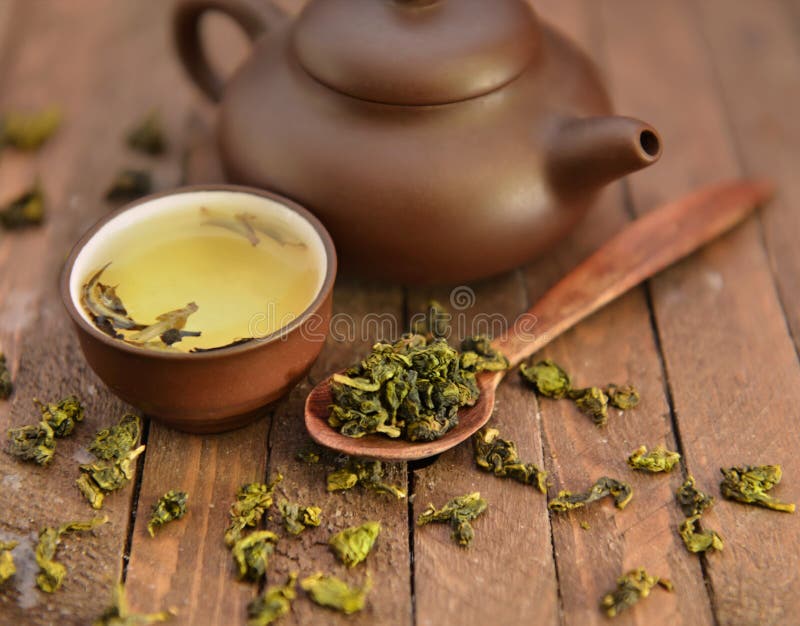 Still life with clay tea pot, small round cup full of fresh green tea and spoon with raw dry tea leaves on wooden background in warm light, asian tea still life. Still life with clay tea pot, small round cup full of fresh green tea and spoon with raw dry tea leaves on wooden background in warm light, asian tea still life