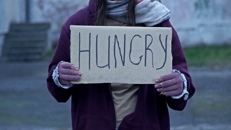 незаметная молодая женщина сидит с попрошайкой и держит написанный рукой голодный плакат.