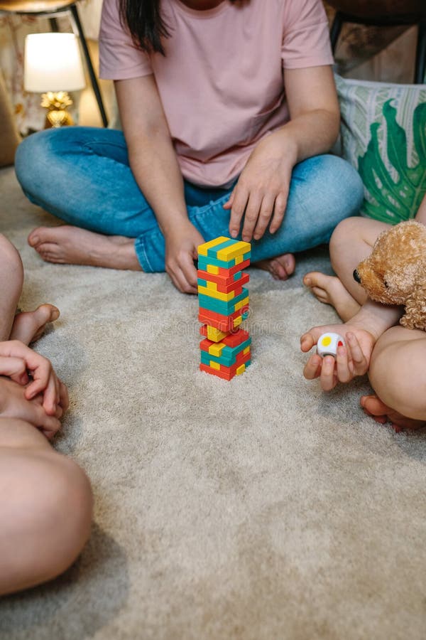 Unrecognizable family having fun making a tower with wooden stacking piece game at home. Mother and sons enjoying together in leisure time. Happy little girl holding a dice and stuffed teddy dog. Unrecognizable family having fun making a tower with wooden stacking piece game at home. Mother and sons enjoying together in leisure time. Happy little girl holding a dice and stuffed teddy dog.